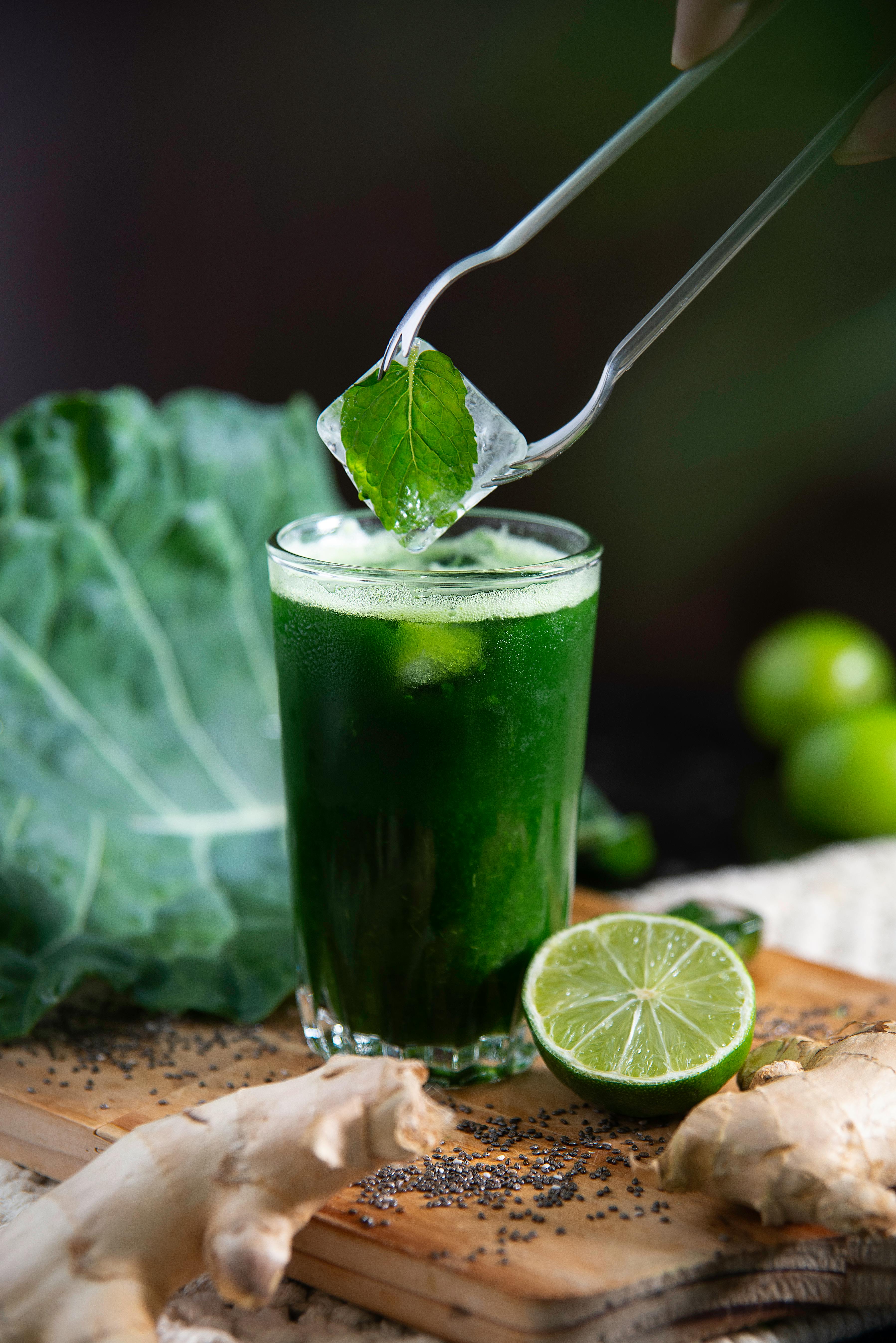 Glass with Green juice, with a green lemon on the side and and ice cube with a mint leaf frozen inside of it being held above it by thongs